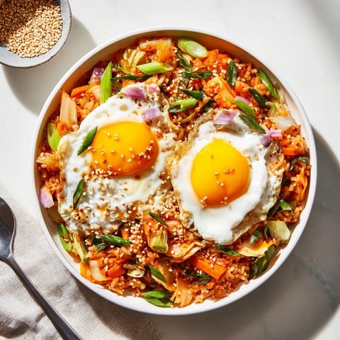 Steam rises from a bowl of kimchi fried rice, studded with diced carrots, onions, and topped with a sunny-side-up fried egg and sesame seeds.